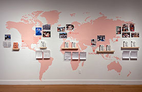 Map of world painted on white wall in pink, with calligraphy and photos calling out New York, Los Angeles, Berlin, Bengaluru, and Tokyo, along with zines on narrow wood shelves.
