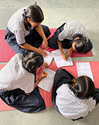Students in school uniforms with braids in loops drawing while seated on the floor on yoga mats.