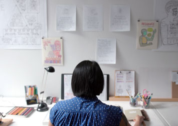 photo of Christine sitting at a desk with drawings on the wall and lots of pencils and art materials on the table