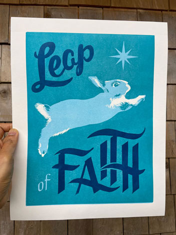 A hand holding a print with calligraphy reading Leap of Faith. A white bunny leaps against a blue green background beneath a light blue star