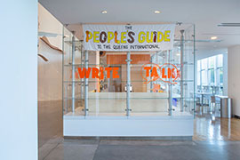 Installation view (exterior of glass-enclosed space) with an embroidered banner with a version of the logo hanging. Inside is pine writing desks. Orange vinyl signage on the glass says write, talk.