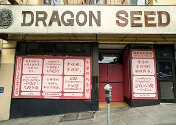 photo of artwork with text in chinese and english in pink and red on a boarded up storefront in Chinatown with masked passersby