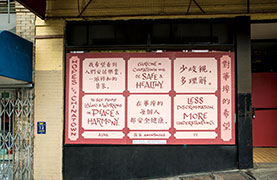 Detail view of the artwork. The text on the poster is in English and Chinese. It reads: Hopes for Chinatown. To see people living and working in peace and harmony, by Alina. Everyone in Chinatown will be safe and healthy. Anonymous. Less discrimination. More Understanding. YY. The text is in red in light pink boxes on a background of red with a scale-like pattern of overlapping concentric circles.