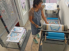 An Asian American woman artist operating a press with a print of a mirror in the drying racks behind her.