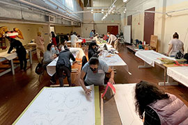 Women working on large tables in an art studio. There are large paper patterns, textiles and tools spread out.