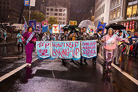 A photo of a contingent with a lead banner in teal and pink with the title of the project in calligraphy, with a mariachi band and banners behind. It's night time and raining but people are smiling.