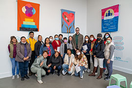 A group photo including many Chinese and Latinx women, one tall white man, and others, in the gallery in front of colorful banners.