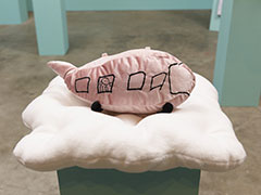 A soft pink stuffed airplane with pom pom wheels resting on a cloud-shaped pillow on a pedestal.