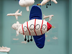 An stuffed airplane with blue wings at the top and bottom of the plane, with a red nose, red stripes and three wheels.