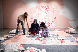 Two adults and one child wearing a onesie assemble the jumbo jigsaw puzzle on a grey carpet.