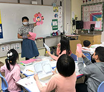 An artist with short hair holds up a sample artwork with a self-portrait in the middle of a paper flower, in front of a classroom with Chinese American elementary school students.