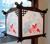 A lantern of wood with fabric that has a papercut in red and blue with a kid pouring cereal into a bowl, a figure in a full moon, and an adult pouring tea for an elder.
