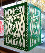 a lantern inspired by papel picado, in green. a woman falls into a safety net. she is clutching a portion of a dollar bill that has been cut by a chain saw
