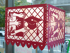 A papel-picado inspired lantern in hot pink showing hands reaching through a door towards a graduation cap
