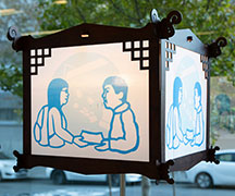 A wooden lantern with a papercut of blues showing two figures interacting in front of an SRO interior scene.