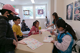 A family with young children collect stamps from two designers inside a gallery, with lanterns in the background