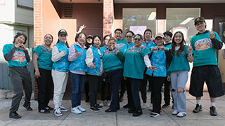 A group photo with a large group of women with a few non-women standing on a sidewalk, smiling and making cute heart-shaped hand gestures.
